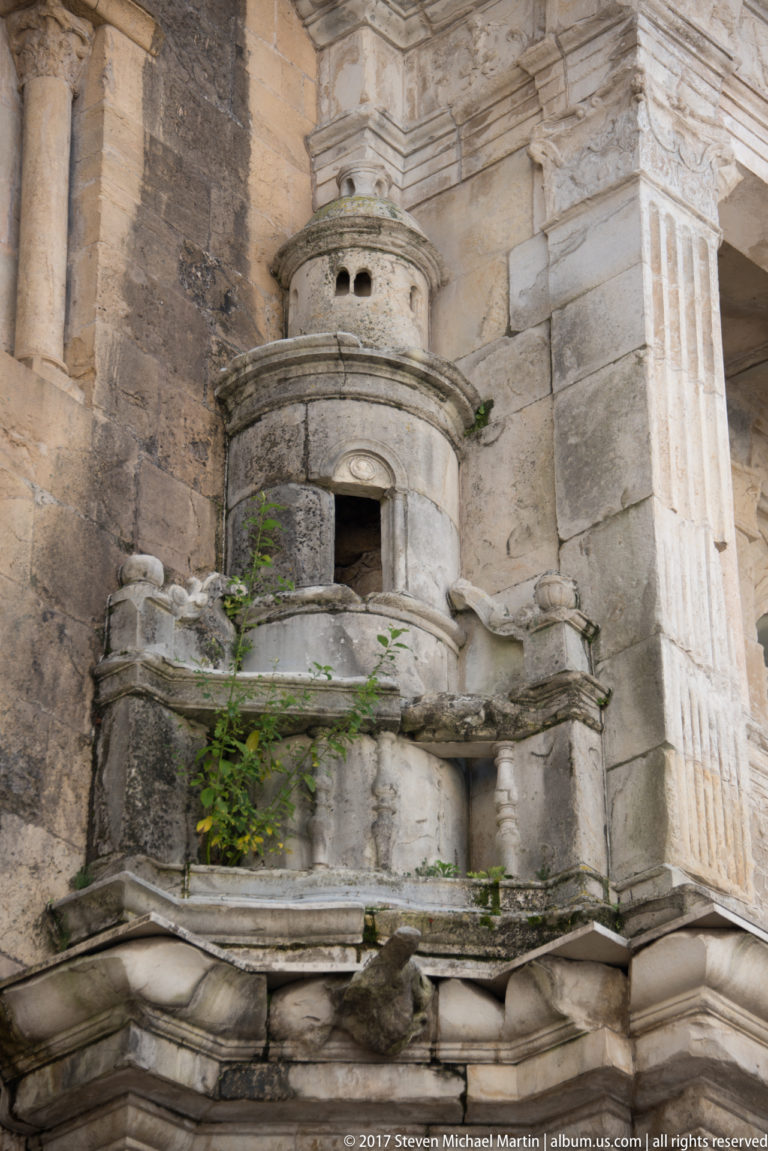 Weathered carved stone architectural detail with small rounded opening, projecting moldings, balustrade, and plants growing from masonry joints.