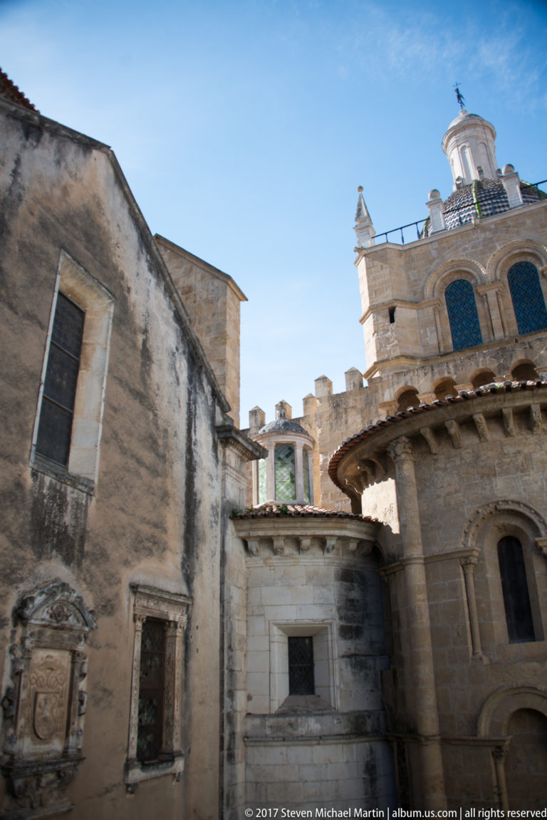 Upward view between weathered stone walls toward crenellations, an ornate upper structure, and a projecting rounded architectural volume.