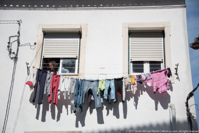 Colorful laundry hangs beneath two shuttered windows on a white wall, casting sharp garment shadows in bright sunlight.