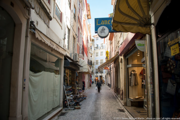 Narrow stone-paved shopping lane with awnings, hanging signs, displays, and pedestrians in Coimbra.