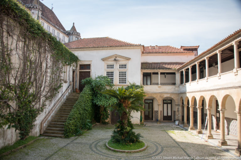 Enclosed whitewashed courtyard with arcaded cloister gallery, stone staircase, patterned paving, tiled roofs, and a circular planted bed.