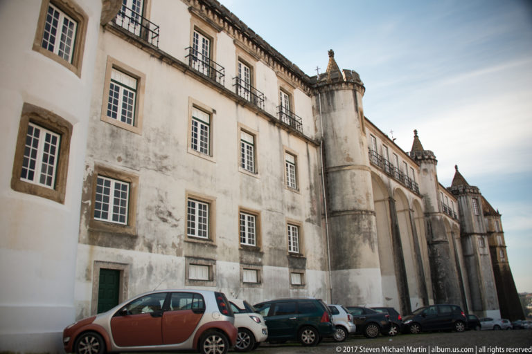 Long white historic building with stone-framed windows, projecting masonry supports, small roofline turrets, and parked cars along its base.