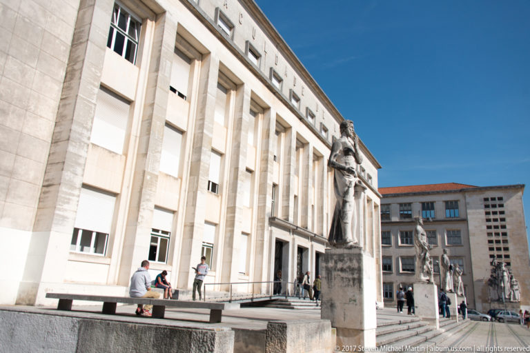 Large pale university building with tall vertical piers, entrance steps, freestanding statues, and people crossing the forecourt.