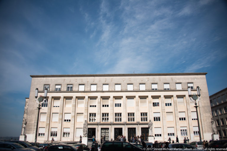 Wide pale academic building with tall vertical window bays, flat parapet, cars, and people beneath a blue sky.