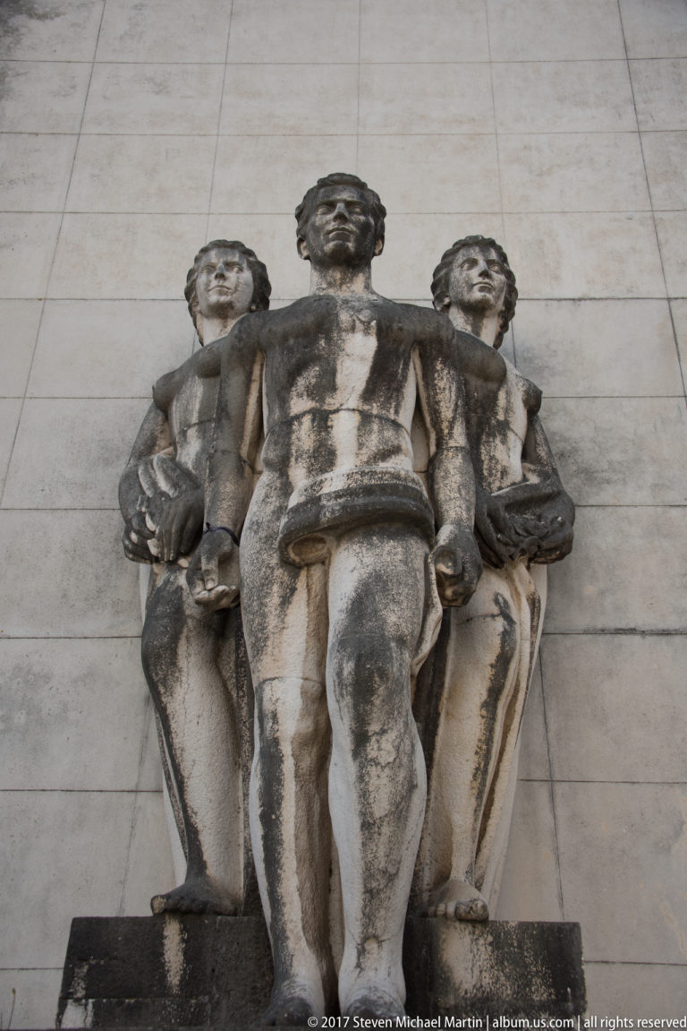 Weathered stone sculpture group of a standing central man flanked by two lower female figures against a pale wall.