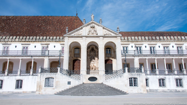 White Via Latina façade with central arched loggia, broad staircase, sculpture, tiled roof, and long galleries in the university courtyard.