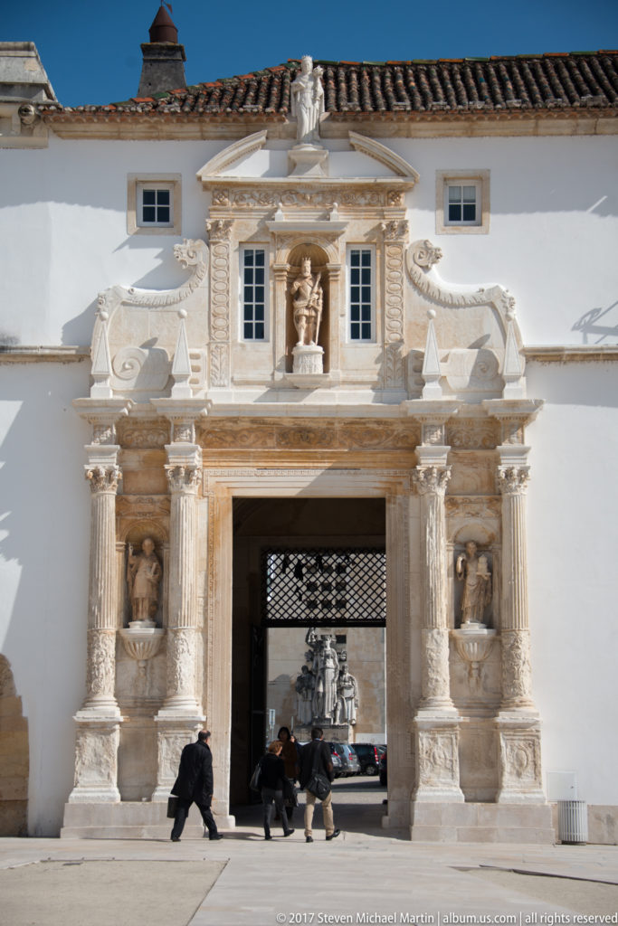 Frontal view of the ornate Porta Férrea with sculpted niches, columns, heraldic ornament, and visitors walking through the central passage.