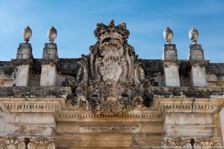 Close view of a crowned royal coat of arms in elaborate weathered Baroque stone carving above the Joanina Library entrance.