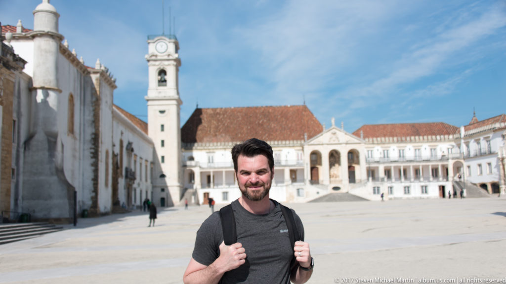 Backpack-wearing visitor stands smiling in the University of Coimbra courtyard, with white palace buildings and the clock tower behind.