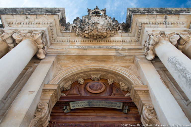 Low-angle detail of the Joanina Library portal showing columns, carved capitals, heraldic crest, curved stonework, and wooden doorway.
