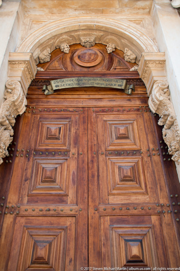 Large paneled wooden double doors with iron studs and fittings beneath the carved stone arch of the Joanina Library portal.