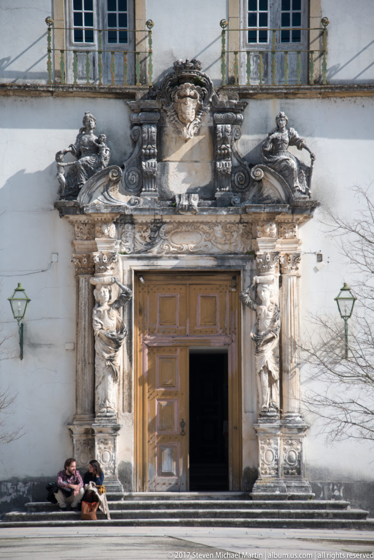 Ornately carved Manueline portal of the Chapel of São Miguel with clustered stonework, heraldic ornament, wooden door, and two seated visitors.