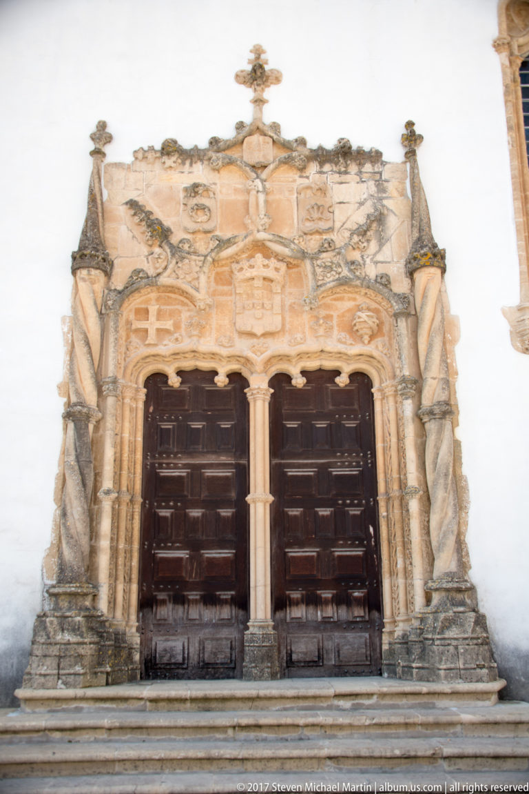 Carved pale-stone double portal with two dark wooden doors, central support, heraldic ornament, pinnacles, and broad entrance steps.