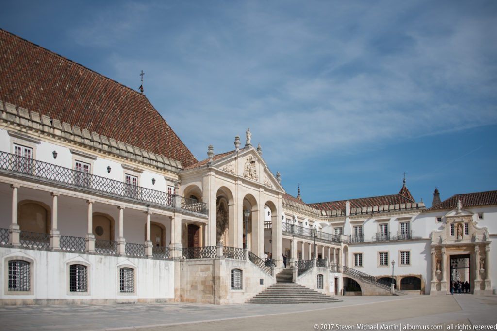 Oblique view across the University of Coimbra courtyard toward the Via Latina, with white galleries, tiled roofs, stairs, and Porta Férrea.