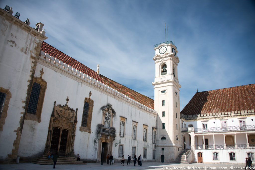 University of Coimbra clock tower rises beside white palace walls, carved doorways, and the tiled-roof Via Latina wing.