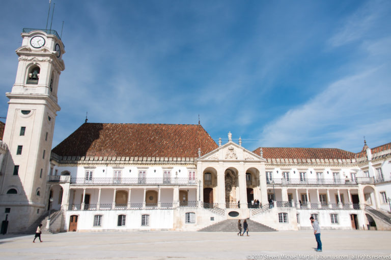 Wide university courtyard with the Via Latina’s central arches and staircase, long white galleries, red tile roof, and clock tower at left.