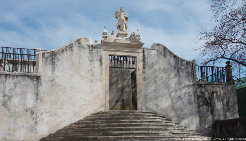 Curved stone steps rise to an iron-gated opening in a weathered white wall crowned by a carved standing figure.
