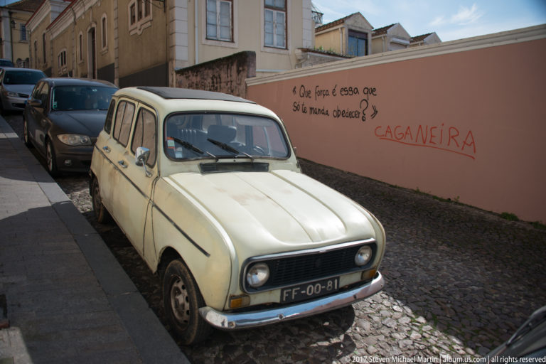 Cream Renault 4 parked on a cobbled street beside a salmon wall with handwritten graffiti and newer cars behind.