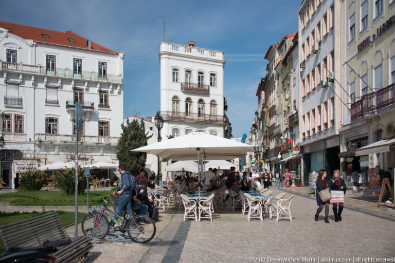 Pedestrians, café seating, bicycles, flower beds, and tall historic buildings fill Largo da Portagem under a bright blue sky.