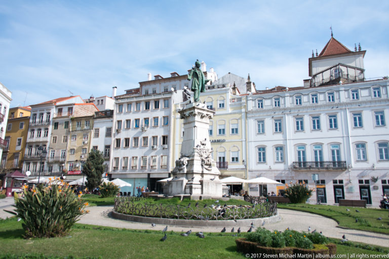 Joaquim António de Aguiar monument rises from a landscaped circle with flowers and pigeons before central Coimbra façades.