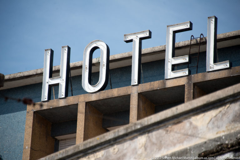 Large white rooftop letters spelling HOTEL rise above concrete bays and a dark roofline against blue sky.