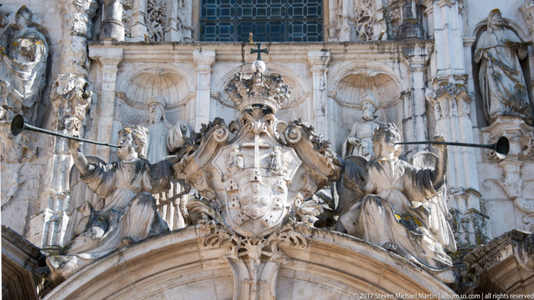 Close view of carved figures, heraldic ornament, scrollwork, and a cross on Santa Cruz Church façade.