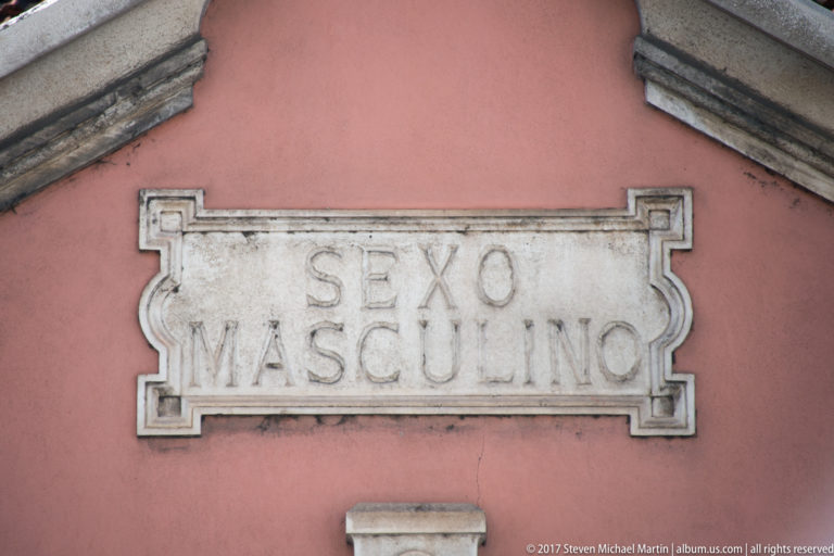 Carved rectangular stone plaque reading “SEXO MASCULINO” mounted on a salmon-colored wall beneath projecting stonework.