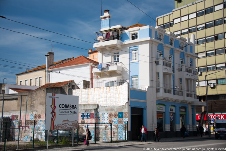 Blue-and-white corner building beside a large “Coimbra é a nossa causa” display, with pedestrians, wires, and mixed urban architecture.
