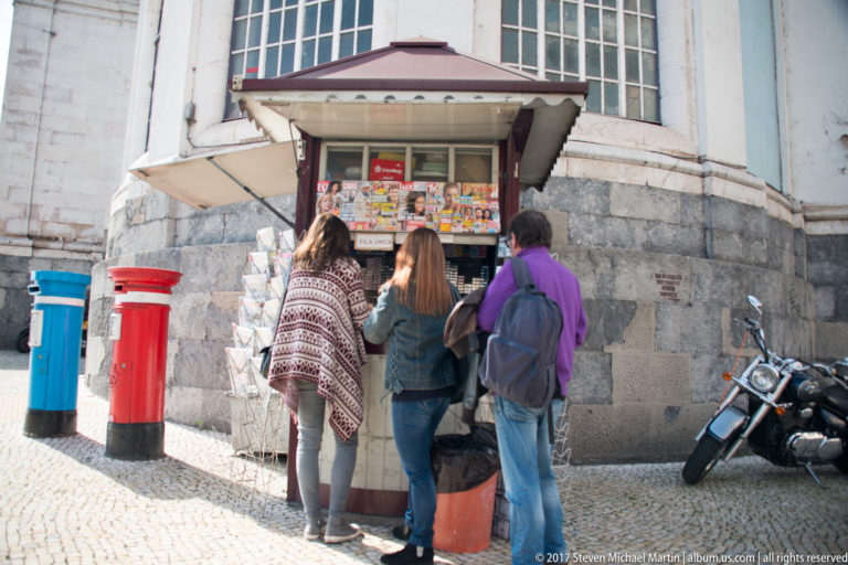 Three women browse a small street kiosk beside red and blue post boxes, with a motorcycle parked against the building.