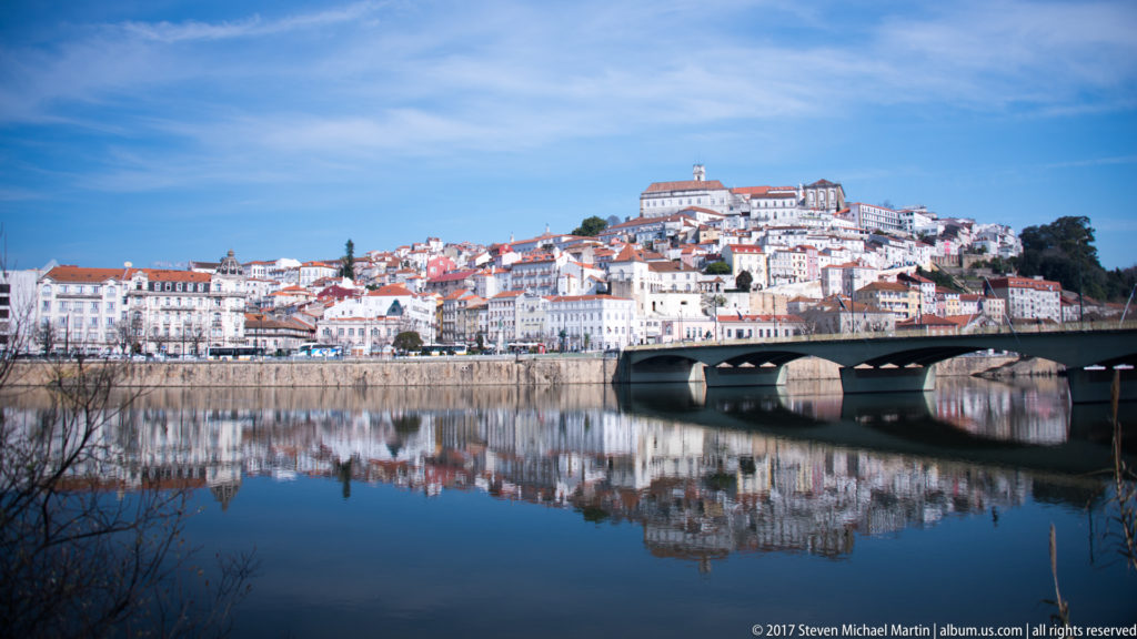 Coimbra hillside and Santa Clara Bridge reflected in calm Mondego River water, with dense red-roofed buildings rising to the hilltop.