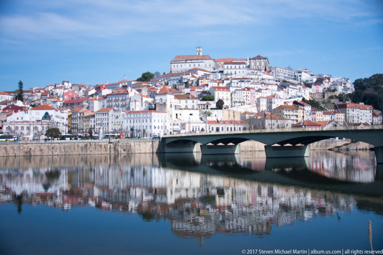 Santa Clara Bridge spans the Mondego before Coimbra’s dense hillside skyline, all mirrored in still river water.