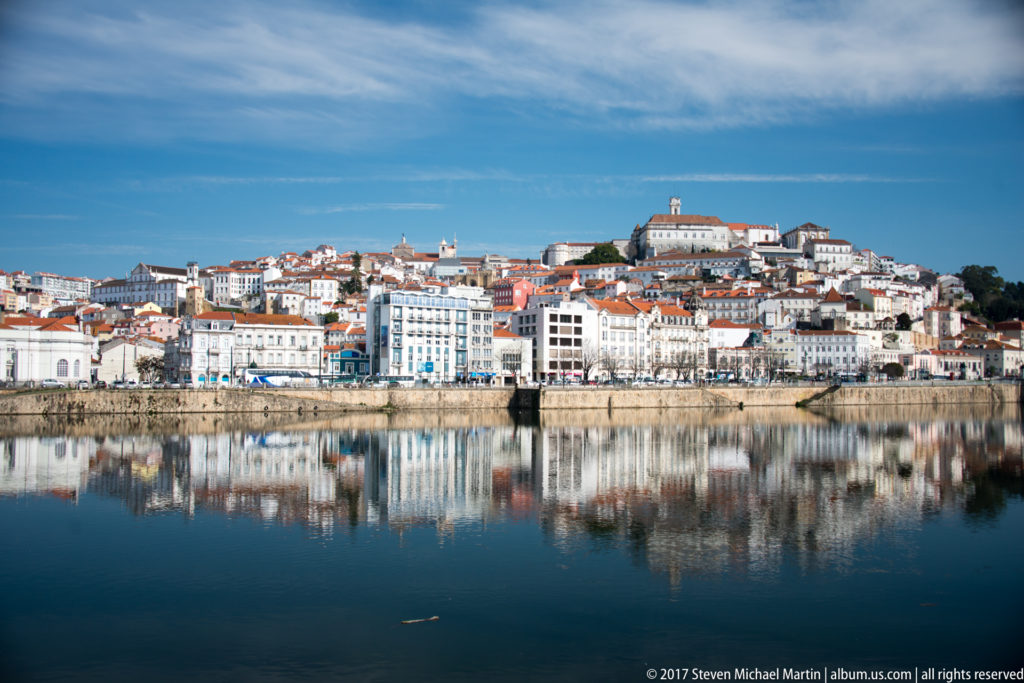 Wide Coimbra skyline of riverfront façades, red-roofed hillside buildings, and hilltop university reflected almost symmetrically in the Mondego.