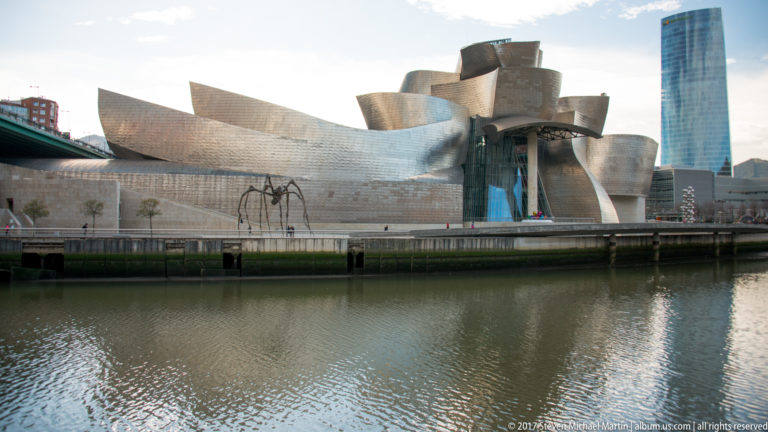 Guggenheim Museum Bilbao across the river, with curving titanium walls, a giant spider sculpture, and a tall glass tower behind.