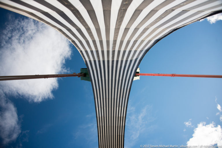 Upward view beneath a flaring striped structural element, crossed by a red beam against blue sky and clouds.