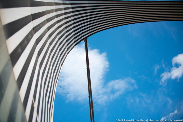Curving black-and-white ribs arc around blue sky while a slender vertical structural member rises through the opening.