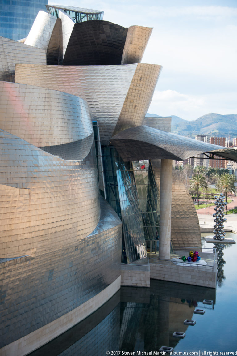 Close oblique view of curved titanium museum walls, glass bay, projecting canopy, water, and reflective sphere sculpture in the distance.