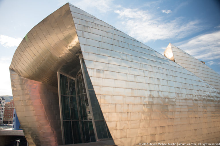 Angular titanium-clad museum volume projects outward above a narrow vertical glass recess under a partly cloudy sky.