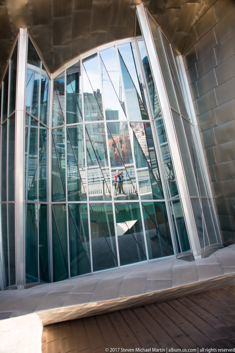 Sloping glass curtain wall with angled metal mullions reflects a bridge and city beneath a titanium-clad overhang.