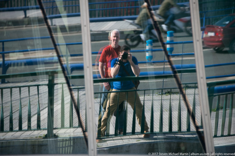 Two people, one holding a camera, reflected in museum glass divided by vertical and diagonal structural lines.