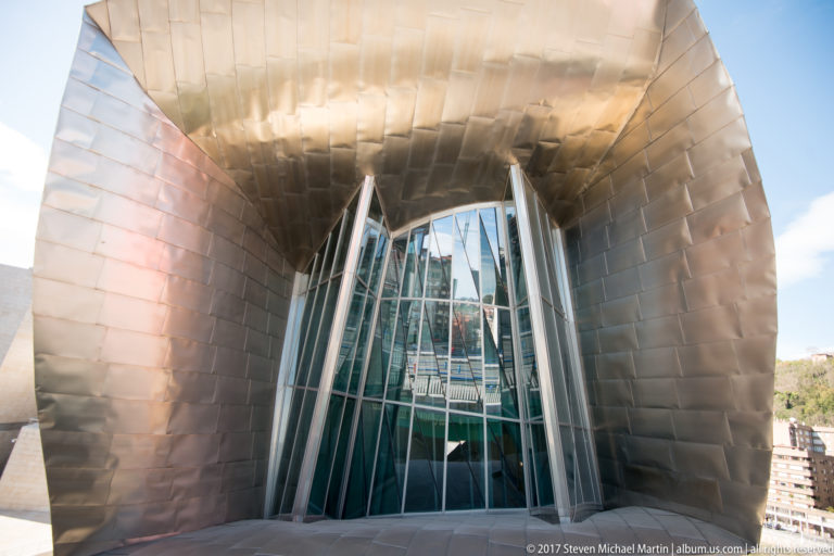 Tall angled glass façade recessed within a deep curving titanium-clad opening at the Guggenheim Museum Bilbao.