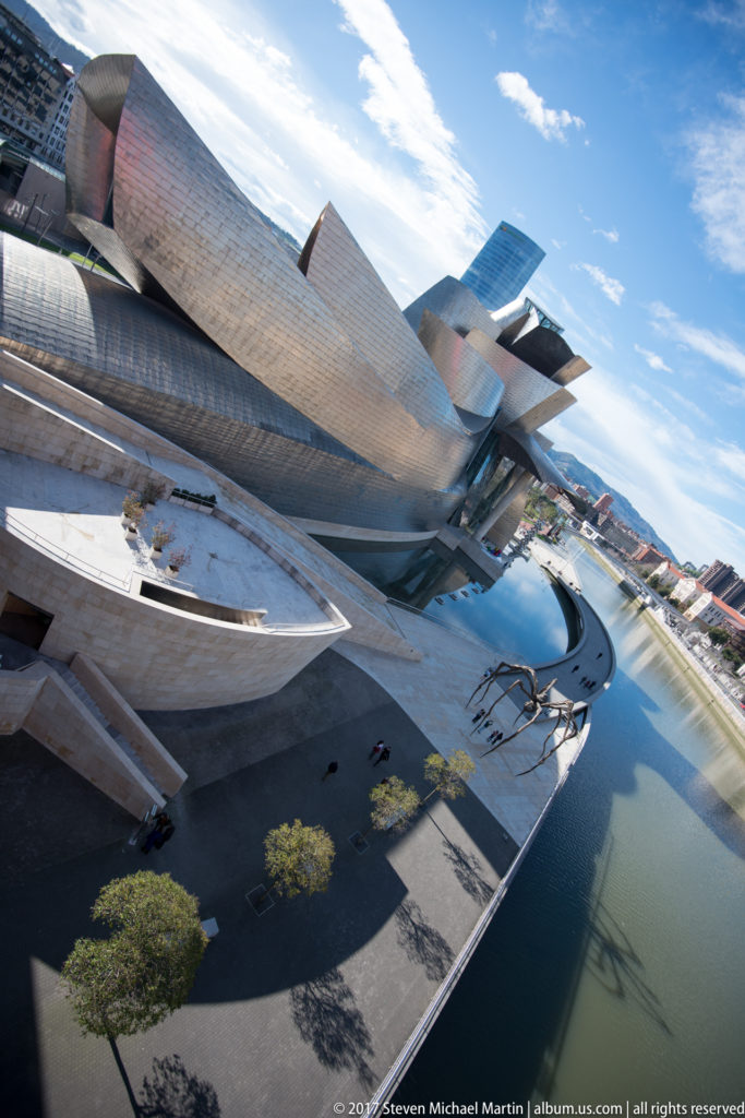 Tilted high-angle panorama of the Guggenheim Museum Bilbao beside the curving river, with Maman and long terrace shadows below.
