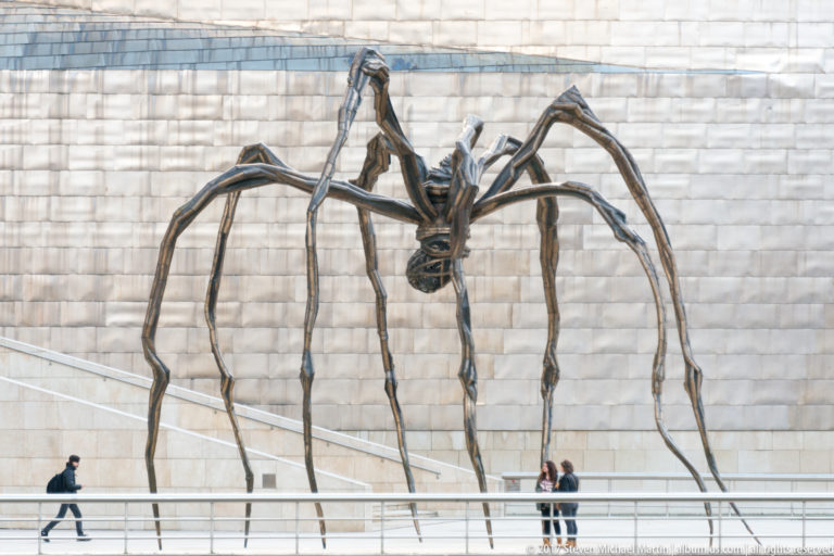 Giant spider sculpture with long bronze legs stands before the museum’s pale metal wall as visitors walk beneath it.