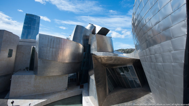 Clustered curved and angular museum volumes surround a recessed glass opening, with a large titanium wall filling the right side.