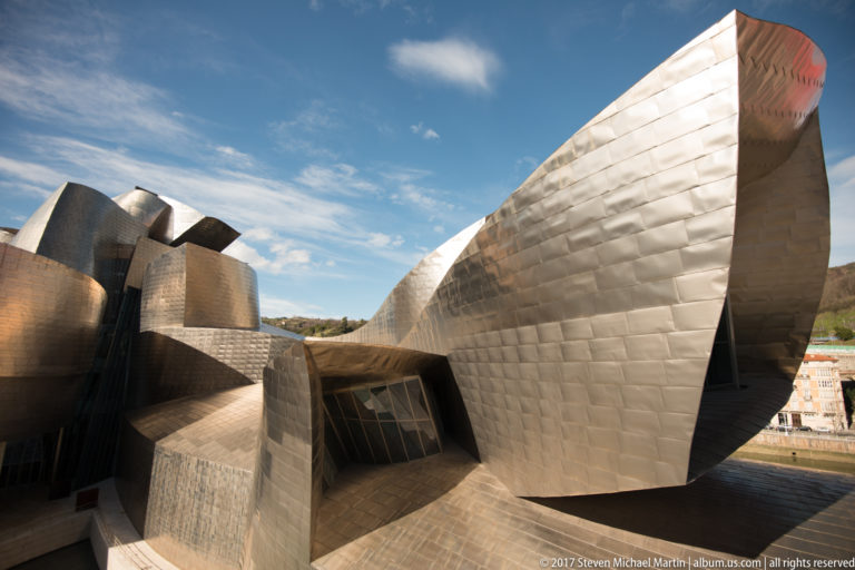 Large angular titanium-clad museum volume projects toward the viewer above recessed glazing, with rounded metal forms behind.