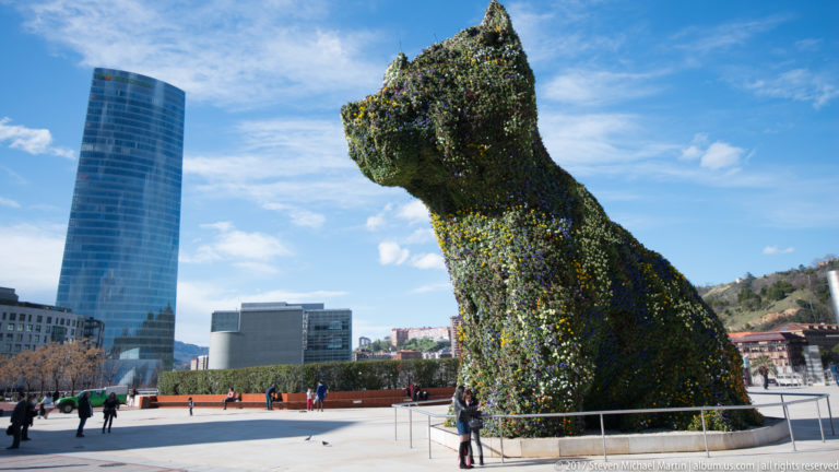 Giant flower-covered terrier sculpture rises above visitors outside the museum, with the Iberdrola Tower at left.