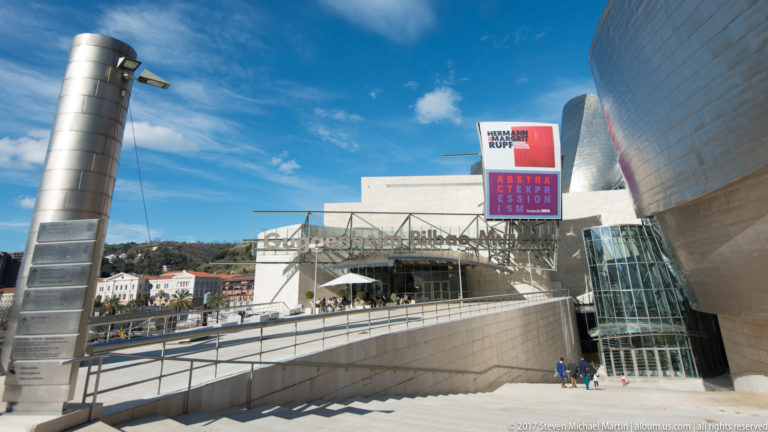 Ramped and stepped approach descends toward the Guggenheim Museum Bilbao entrance, with glass walls, signage, and an exhibition banner.