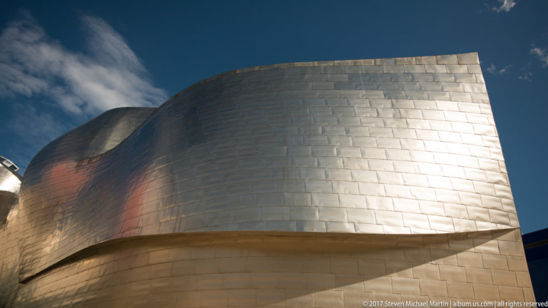 Close view of a broad curved titanium-clad museum wall above pale stone, catching bright sunlight under deep blue sky.