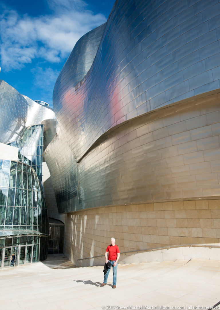 Visitor stands beneath sweeping titanium museum walls beside a faceted glass entrance, with sharp shadows across the pale pavement.