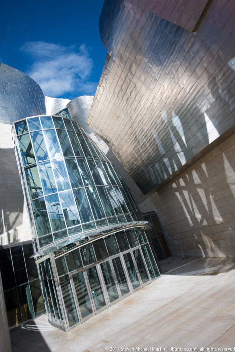 Faceted glass entrance with angled steel mullions sits beneath an overhanging titanium museum form casting geometric shadows.