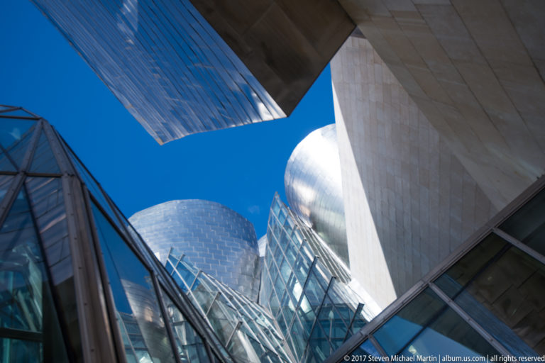 Upward view between glass, stone, and titanium museum volumes framing a wedge of vivid blue sky.